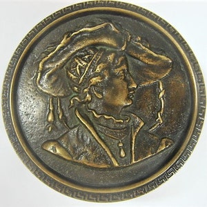 May include: A bronze-toned, circular relief sculpture featuring a profile of a person wearing a hat. The figure is set within a decorative border. The sculpture has a classical aesthetic, with intricate details and a weathered appearance, suggesting an antique or vintage piece.