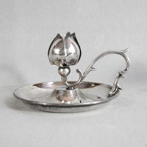 May include: A silver candlestick holder with a tulip-shaped top and a round base. The handle curves gracefully, adding to the elegant design. The candlestick holder is made of a shiny, reflective metal.