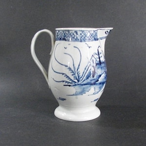 May include: White ceramic pitcher with a blue landscape design. The pitcher features a curved handle and a spout for pouring. The blue design includes a house, trees, and water, with decorative patterns around the rim.
