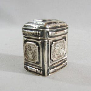 May include: A small, silver-colored box with a rectangular shape and ornate detailing. The box features decorative panels with embossed designs and a hinged lid. The overall design suggests a vintage or antique style, with a polished finish.