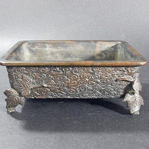 Puede incluir: Un macetero cuadrado de bronce con un patrón decorativo de nubes en espiral y cuatro patas en forma de hoja.