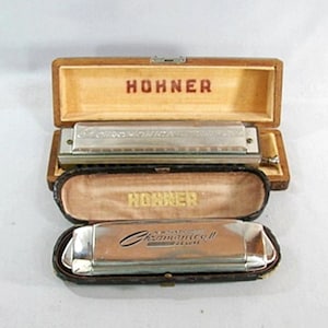 May include: Two Hohner harmonicas in their original cases. The top case is open, revealing a silver harmonica with the text "Echo" and "Hohner" in red. The bottom case is also open, displaying a silver harmonica with the text "Romantic II Deluxe".