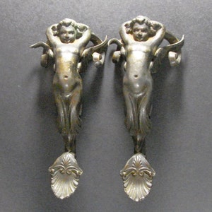 May include: A pair of antique silver-toned metal door pulls shaped like cherubs with outstretched wings. The cherubs are holding onto a scroll-like design.