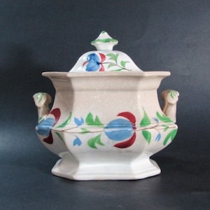 May include: A white porcelain sugar bowl with a lid, decorated with hand-painted floral designs in shades of blue, red, and green. The bowl has a scalloped edge and two handles.