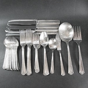 May include: A collection of silver-colored flatware, including knives, forks, spoons, and serving spoons, arranged on a dark surface. The silverware has a classic design, suitable for formal dining.