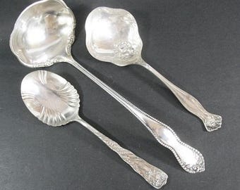May include: Three ornate silver serving spoons and ladles. The spoons have decorative handles and bowl shapes. The ladle has a deep, scalloped bowl. The items are arranged on a dark surface.