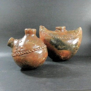 May include: Two brown ceramic pottery jugs with decorative details. The jug on the left has a round body and a spout, while the jug on the right has a curved body.