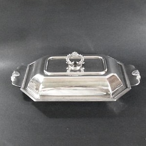 May include: A silver-colored, rectangular serving dish with a lid. The dish has a decorative handle on the lid and ornate detailing on the sides. The dish is likely made of metal and is designed for serving food.