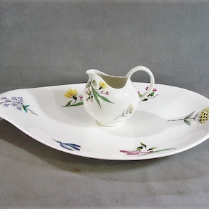 May include: A white ceramic serving dish with a matching creamer. The dish has a unique, elongated shape and is decorated with colorful floral designs in yellow, purple, and pink. The creamer sits in the center of the dish and also features floral accents.