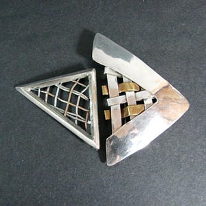 May include: A silver-toned geometric brooch with a triangular section featuring a woven pattern of silver and gold-toned metal. The other section is a silver-toned, asymmetrical shape with a woven pattern of silver and gold-toned metal.