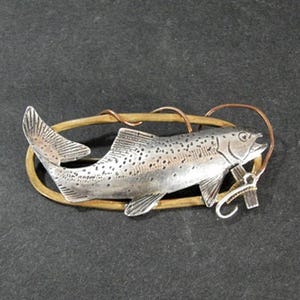May include: A silver-toned fish brooch with a detailed design, featuring a fish with a hook and line. The brooch has a gold-colored oval frame and copper-colored accents, perfect for fishing enthusiasts.