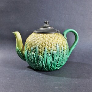 May include: A ceramic teapot featuring a textured pineapple-inspired body with a yellow upper section and green leaf-like base. The teapot includes a curved green handle, a matching spout, and a dark metallic lid topped with a small finial.