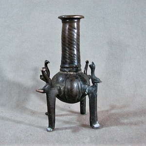 May include: A dark brown metal vase with a twisted neck and three legs shaped like animal heads. The vase has a round body and a textured surface.