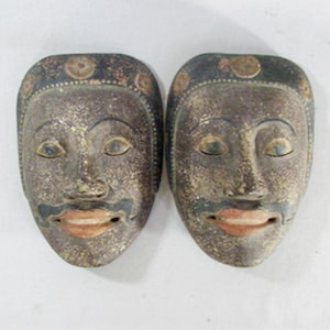 May include: Two antique wooden masks with a weathered appearance. Each mask features a stylized face with a dark gray head, a black mustache, and a red-orange mouth. The masks have a traditional design, possibly for cultural or decorative purposes.