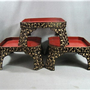 May include: Three black lacquered wooden trays with gold leaf floral designs. The trays are stacked on top of each other and have red interiors.