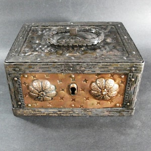 May include: A rectangular metal box with a hinged lid and a handle. The box has a silver-colored top and sides, with a copper-colored front panel. The front panel features two floral designs and a keyhole.