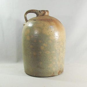 May include: A stoneware jug with a rounded body and a handle. The jug has a mottled, speckled appearance with shades of tan, brown, and gray. The jug is likely an antique or vintage piece, suitable for rustic decor.