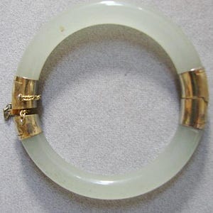 May include: A circular bangle bracelet featuring a smooth, translucent pale green stone band. The bracelet is accented with two polished gold-toned metal segments, one of which includes a hinged clasp mechanism and a small safety chain attachment.