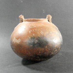 May include: A round, earthenware pot with a reddish-brown exterior and dark patches. The pot has two small, decorative handles near the rim. The surface shows signs of wear, adding to its rustic charm.