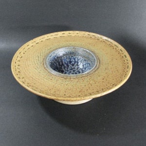 May include: A ceramic bowl with a wide, flat base and a smaller, round bowl in the center. The base is a light brown color with a textured surface. The smaller bowl is a dark blue color with a white and blue speckled pattern.