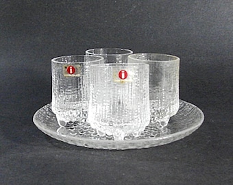 May include: A set of four clear textured glass tumblers with a matching round tray. Each tumbler has a red and white sticker. The glassware is designed with vertical lines and sits on a clear, textured tray.