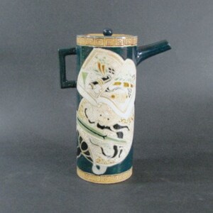 May include: A tall, cylindrical ceramic pitcher with a green glaze and a white and gold design. The pitcher has a spout and a handle.