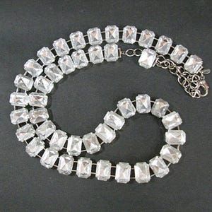 May include: A silver-toned necklace featuring rectangular, clear crystal-like stones. The stones are set in a row, connected by a silver chain. The necklace has a clasp closure and is displayed on a dark background.