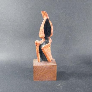May include: Abstract wooden sculpture of a person in a seated pose, painted in black and orange hues, mounted on a wooden base.