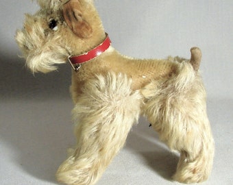 May include: A plush toy dog with a red collar. The dog is light brown and has a fluffy, shaggy coat. The dog is standing on a white surface.
