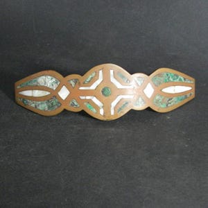 May include: A copper-toned barrette with inlaid designs. The barrette features a central cross-like pattern with white and green accents, and oval-shaped sections with green and white inlays on each end. The barrette is set against a dark background.