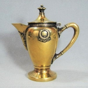 May include: A gold-tone metal creamer or pitcher with a hinged lid and a decorative handle. The creamer has a detailed design with a crest on the front.