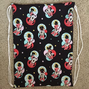 K&ouml;nnte beinhalten: Schwarze Kordelzugtasche mit einem sich wiederholenden Muster von Minnie Mouse in Rot, Wei&szlig; und Gelb auf schwarzem Hintergrund. Die Tasche hat wei&szlig;e Kordeln.