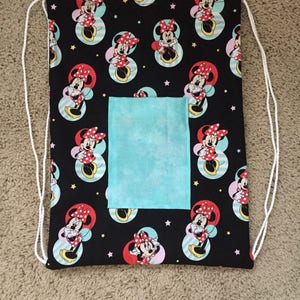 K&ouml;nnte beinhalten: Eine schwarze Kordeltasche mit einem Minnie Mouse-Druck. Die Tasche hat eine kleine, hellblaue Tasche auf der Vorderseite.