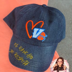 Op de afbeelding: Denim baseballpet met een geborduurd ontwerp. Het ontwerp bevat een hart, de letter "V" en een basketbal in oranje en blauw. De tekst "El Rancho Se Respeta" is met goudkleurig garen op de rand geborduurd.
