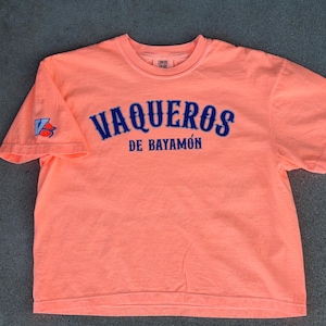 May include: Coral t-shirt featuring the text "VAQUEROS DE BAYAMÓN" in blue arched lettering. A blue and white logo is on the left sleeve. The short-sleeved shirt is made of a soft material.