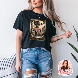 Op de afbeelding: Zwart T-shirt met een ingelijste afbeelding van een skelet in een roze shirt, met een rode hoofddoek en een vliegtuig. De tekst "THE MILITARY SPOUSE" staat eronder. Het T-shirt wordt gedragen met een blauwe jeans.