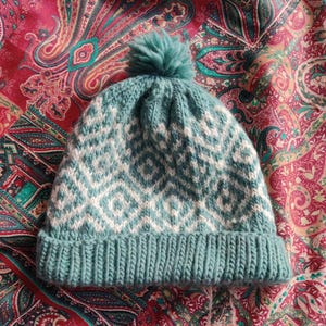 Puede incluir: Un gorro de punto azul turquesa con un patrón geométrico blanco y un pompón esponjoso en la parte superior.