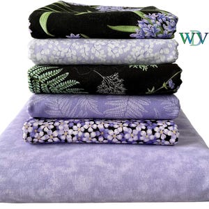 May include: A stack of six folded fabric pieces featuring various floral and botanical patterns in shades of purple, black, and white. The top piece is black with purple flowers, followed by light purple with white leaves, black with green ferns, light purple with fern patterns, purple flowers on black, and a solid light purple base.