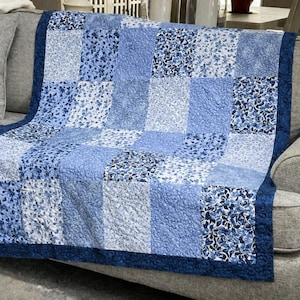 May include: A patchwork quilt featuring a repeating grid of square fabric panels in various shades of blue and white. The design alternates between solid light blue textures and intricate floral patterns, all framed by a dark blue border.