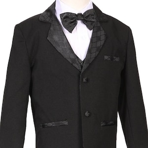 May include: A black tuxedo suit for a young boy. The suit includes a jacket, vest, and bow tie. The jacket has a satin lapel with a diamond pattern. The vest has a similar diamond pattern. The bow tie is black satin.