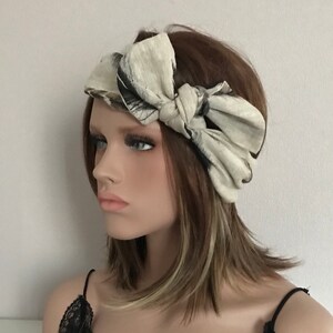 May include: A cream-colored headband with a large bow, featuring black and gray abstract patterns. The headband is tied in a knot at the top, creating a stylish accessory. The fabric has a textured appearance, adding visual interest.