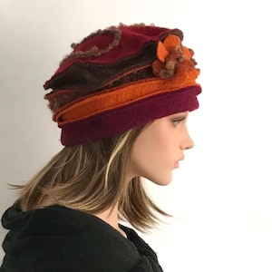 Puede incluir: Un sombrero de fieltro con bandas superpuestas en color burdeos intenso, marrón oscuro y naranja brillante. El sombrero está adornado en el lateral con un grupo de detalles florales de fieltro circulares y texturizados en tonos naranja y marrón.