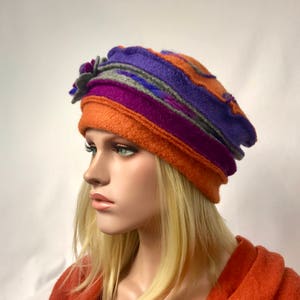 Peut inclure: Un chapeau en feutre coloré orné d'une fleur. Le chapeau présente des bandes orange, violet, grise et magenta. Le chapeau est porté par un mannequin aux cheveux blonds et une veste orange.