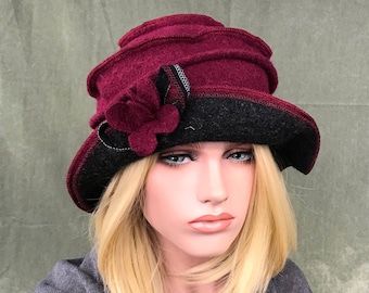 May include: A burgundy felt hat with a black felt brim and a burgundy felt flower accent. The hat has a layered design and a slightly slouched crown.