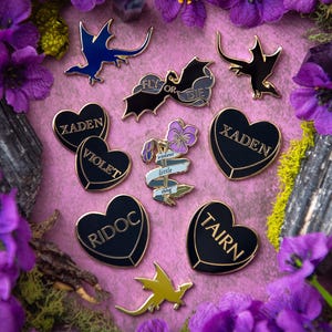 Könnte beinhalten: Eine Sammlung von Emaille-Pins mit Drachen, Herzen und einer Blume mit dem Text "Fly or Die", "Xaden", "Violet", "Ridoc", "Tairn" und "violent little thing". Die Pins sind auf einem rosa Hintergrund angeordnet, der von lila Blumen umgeben ist.