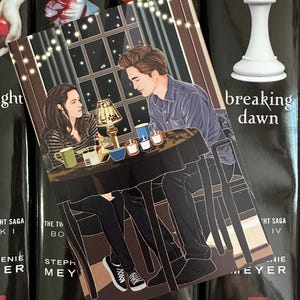 以下が含まれることがあります： テーブルに座っている2人のイラストが描かれたブックマーク。背景には窓とストリングライトが描かれています。ブックマークは「Breaking Dawn」というタイトルの本の上に置かれています。ブックマークには「MT Books」と「LITTLE, BROWN」の文字があります。