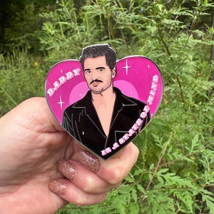 May include: A heart-shaped pin with a portrait of a man in a black shirt, set against a pink background. The pin features the text "DADDY" and "in a state of mind". The pin is held in a hand with glittery nail polish.