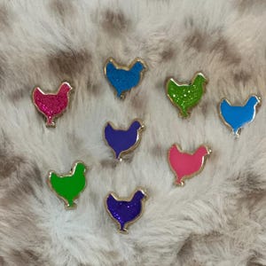May include: Eight colourful glitter chicken pins in various colours including blue, green, pink, and purple. The pins are all the same shape and size and are arranged in a semi-circle on a white fluffy surface.