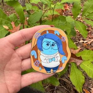 May include: A colorful enamel pin featuring the character Sadness from the movie Inside Out. The pin shows Sadness with blue hair, a white sweater, and blue jeans. The pin has a tan border with blue tear drop accents.