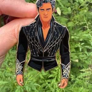 May include: Black enamel pin featuring a stylized male figure with orange skin, dark hair, and a black outfit with gold accents. The pin has the text "The High Lord arrived" on a black background.